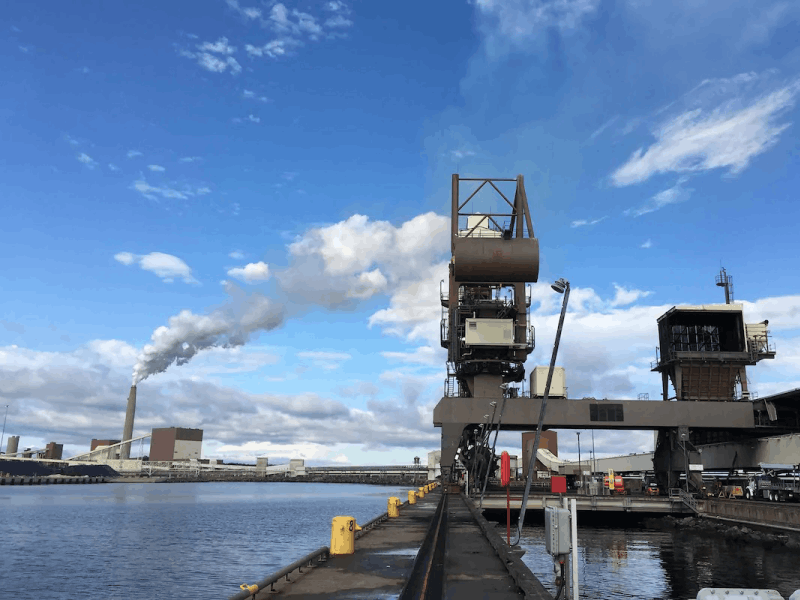 Industrial waterfront with cargo-handling cranes, heavy machinery, and a smokestack emitting a visible plume, illustrating the emissions challenges facing ports and heavy industry and the need for technologies that reduce harmful pollutants and improve environmental performance.
